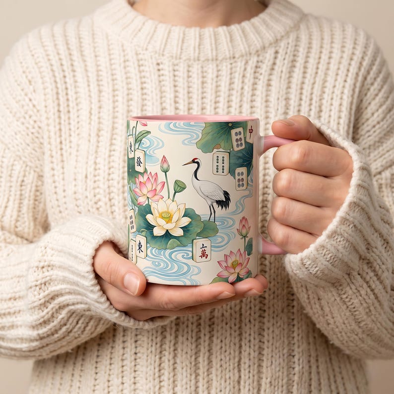 May include: A ceramic mug with a light pink interior and handle, featuring a detailed illustration of a white crane, pink lotus flowers, green lily pads, blue water ripples, and various mahjong tiles with characters like 東, 發, and 中.