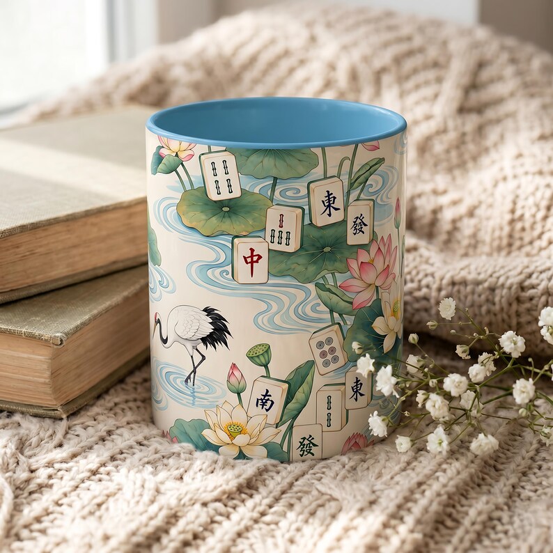 May include: A ceramic mug with a light blue interior and a cream-colored exterior featuring a pattern of lotus flowers, green lily pads, a white crane, and various mahjong tiles with Chinese characters and symbols printed across the surface.