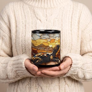 May include: A black ceramic mug featuring a wrap-around stained glass style design. The artwork depicts a sea turtle in shades of blue, brown, and gold, set against a background of abstract geometric shapes in warm yellow and amber tones.
