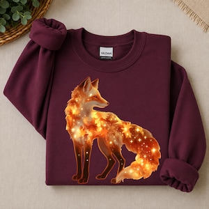 Könnte beinhalten: Ein kastanienbraunes Langarm-Sweatshirt mit einer Grafik eines Fuchses, der aus leuchtenden orangefarbenen und gelben Herbstblättern sowie kleinen weißen Sternen besteht. Das Gildan-Markenetikett ist am inneren Rundhalsausschnitt sichtbar.