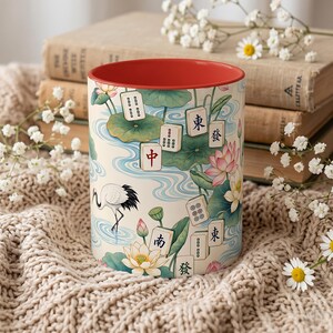 May include: A ceramic mug with a red interior and a cream-colored exterior featuring a pattern of Mahjong tiles, pink lotus flowers, green lily pads, and a white crane. The tiles display various Chinese characters and symbols in black and red.