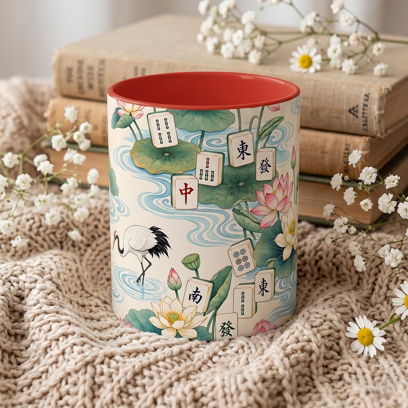 May include: A ceramic mug with a red interior and a cream-colored exterior featuring a pattern of Mahjong tiles, pink lotus flowers, green lily pads, and a white crane. The tiles display various Chinese characters and symbols in black and red.