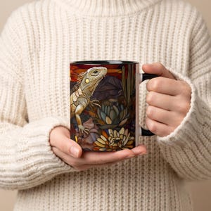 Puede incluir: Una taza de cerámica con una ilustración estilo vidriera que muestra una iguana rodeada de flora desértica, incluyendo cactus y suculentas. El diseño presenta tonos cálidos en naranja, marrón y verde, con un asa y borde interior negros.