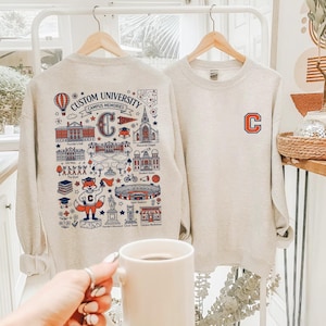 May include: Two light gray sweatshirts hang on wooden hangers. One features a large graphic with the text "CUSTOM UNIVERSITY CAMPUS MEMORIES" and a fox mascot. The other has a large "C" on the front.