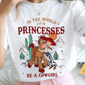 Puede incluir: Camiseta blanca con un gr&aacute;fico de dibujos animados. El texto dice "IN THE WORLD full of PRINCESSES BE A COWGIRL". El gr&aacute;fico muestra una vaquera, un caballo, cactus y un paisaje des&eacute;rtico. Una prenda casual.