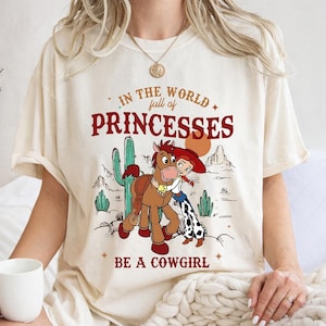 Puede incluir: Camiseta color crema con un gr&aacute;fico que presenta a una vaquera de dibujos animados abrazando a un caballo, sobre un paisaje des&eacute;rtico con cactus y monta&ntilde;as. El texto dice: "In the world full of princesses, be a cowgirl."