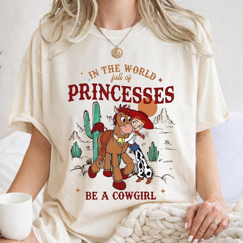 Puede incluir: Camiseta color crema con un gr&aacute;fico que presenta a una vaquera de dibujos animados abrazando a un caballo, sobre un paisaje des&eacute;rtico con cactus y monta&ntilde;as. El texto dice: "In the world full of princesses, be a cowgirl."