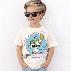 Può includere: Una maglietta color crema con un'immagine a cartoni animati di Buzz Lightyear che tiene in mano un boccale di birra. La maglietta include il testo "Buzz Lite Beer Star Command Brewery Since 1995". La maglietta è indossata da una persona giovane con occhiali da sole neri.