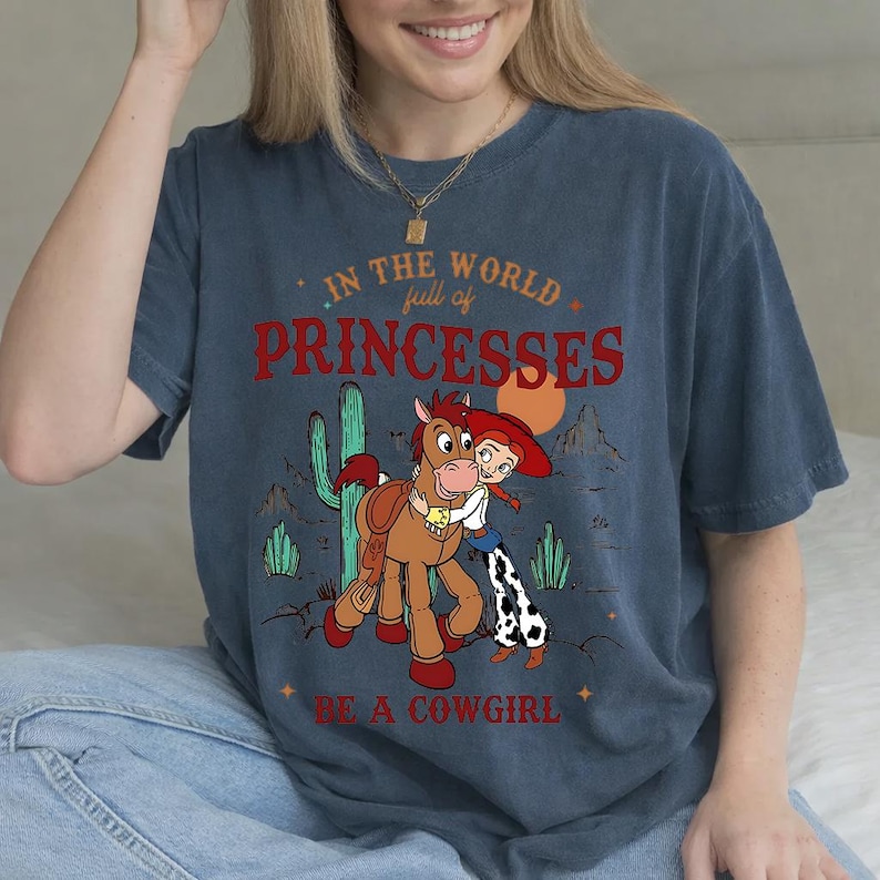 Puede incluir: Camiseta azul marino con un dise&ntilde;o de vaquera de dibujos animados y un caballo, sobre un fondo de paisaje des&eacute;rtico. El texto de la camiseta dice: "In the world full of princesses, be a cowgirl."