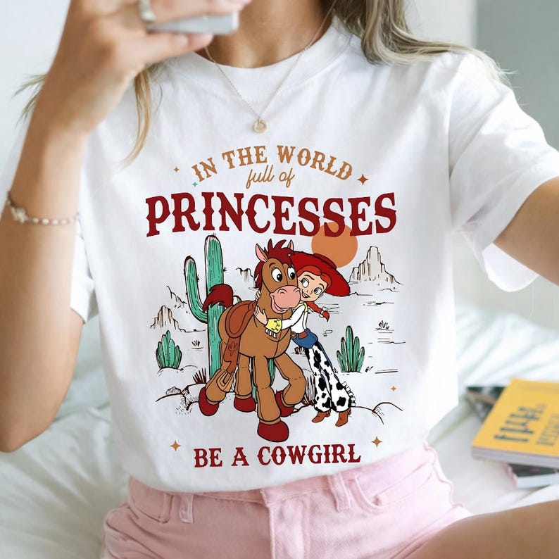 Puede incluir: Camiseta blanca con un gr&aacute;fico de dibujos animados. El texto dice "IN THE WORLD full of PRINCESSES BE A COWGIRL". El gr&aacute;fico muestra una vaquera, un caballo, cactus y un paisaje des&eacute;rtico. Una prenda casual.