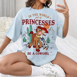 Puede incluir: Camiseta azul claro con un gr&aacute;fico de dibujos animados de una vaquera y un caballo, con el texto "IN THE WORLD full of PRINCESSES BE A COWGIRL". El dise&ntilde;o incluye cactus y un paisaje des&eacute;rtico.