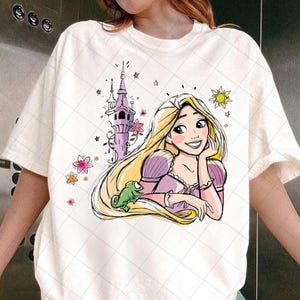 May include: White t-shirt featuring a cartoon illustration of a princess with long blonde hair, a purple dress, and a green chameleon. A purple tower, stars, and flowers are also depicted in the design.
