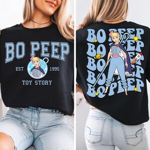 Può includere: Felpa nera con un design di Bo Peep di Toy Story. La parte anteriore mostra "BO PEEP" in azzurro, con un'illustrazione di Bo Peep e il testo "EST. 1995 TOY STORY." Il retro presenta una grafica più grande di Bo Peep con testo.