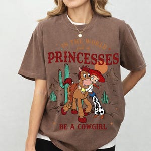 Puede incluir: Camiseta marr&oacute;n con un dibujo animado de una vaquera y un caballo en un paisaje des&eacute;rtico. El texto dice: "En el mundo lleno de princesas, s&eacute; una vaquera."