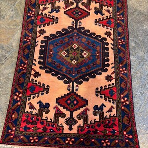 May include: A rectangular, hand-woven rug with a central peach-colored field. The rug features a detailed geometric pattern in shades of navy blue, red, and brown. The border is a deep navy blue with repeating floral motifs. The rug's design is symmetrical.