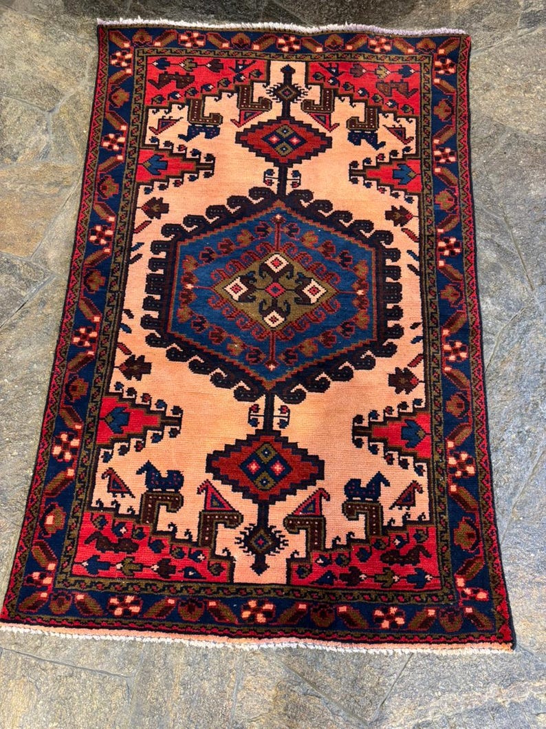 May include: A rectangular, hand-woven rug with a central peach-colored field. The rug features a detailed geometric pattern in shades of navy blue, red, and brown. The border is a deep navy blue with repeating floral motifs. The rug's design is symmetrical.