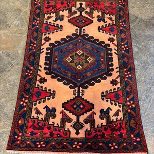 May include: A rectangular rug with a central peach-colored field and intricate geometric patterns. The rug features a navy blue border with red and brown accents. The central design includes a large blue diamond shape and smaller red and brown motifs.