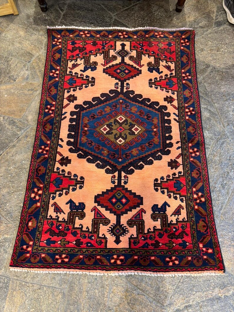 May include: A rectangular rug with a central peach-colored field and intricate geometric patterns. The rug features a navy blue border with red and brown accents. The central design includes a large blue diamond shape and smaller red and brown motifs.