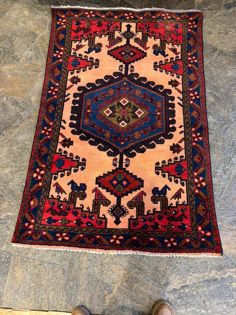 May include: A rectangular rug with a central peach field, framed by a navy blue and red border. The rug features geometric patterns in navy, red, and brown, with a central diamond-shaped design. The rug's dimensions are approximately 3 x 5 feet.