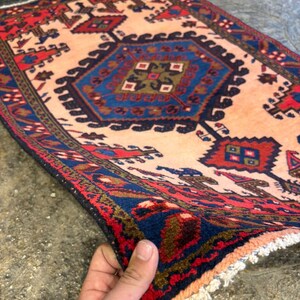 May include: A rectangular rug with a central geometric design in shades of blue, red, and brown on a beige background. The rug has a detailed border and fringe. The rug is being held up, showing its texture and pattern.