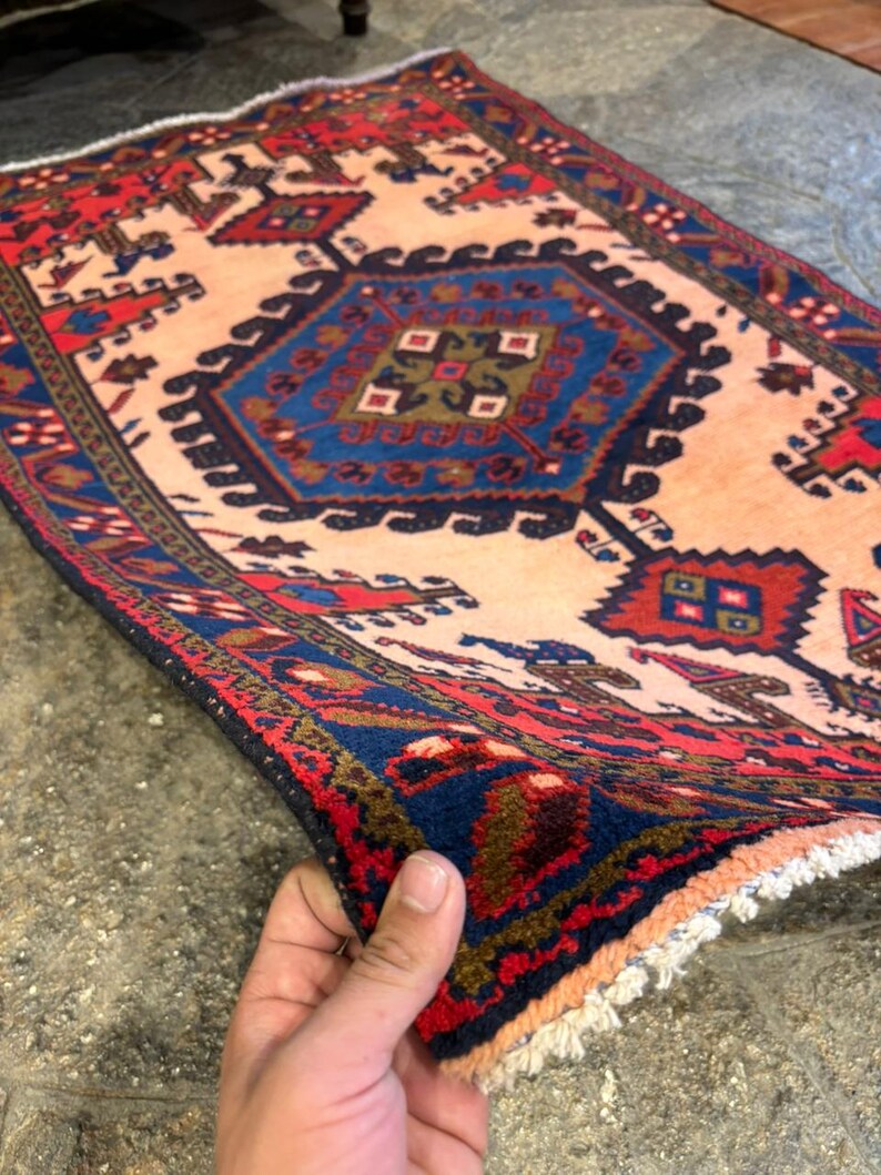May include: A rectangular rug with a central geometric design in shades of blue, red, and brown on a beige background. The rug has a detailed border and fringe. The rug is being held up, showing its texture and pattern.