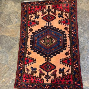 May include: Rectangular rug with a central geometric design in shades of blue, red, and brown on a peach background. The rug has a detailed border with repeating patterns and a fringed edge. The rug's design is symmetrical and traditional.
