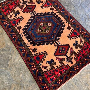 May include: Rectangular rug with a central medallion in shades of blue, brown, and green, set against a peach-colored field. The rug features a border of navy blue and red, with intricate geometric patterns. The rug is likely made of wool.