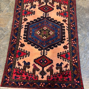 May include: A rectangular rug with a central peach-colored field and intricate geometric patterns. The border is a deep navy blue with red and brown accents. The rug features repeating motifs of stylized shapes and figures, creating a traditional aesthetic.