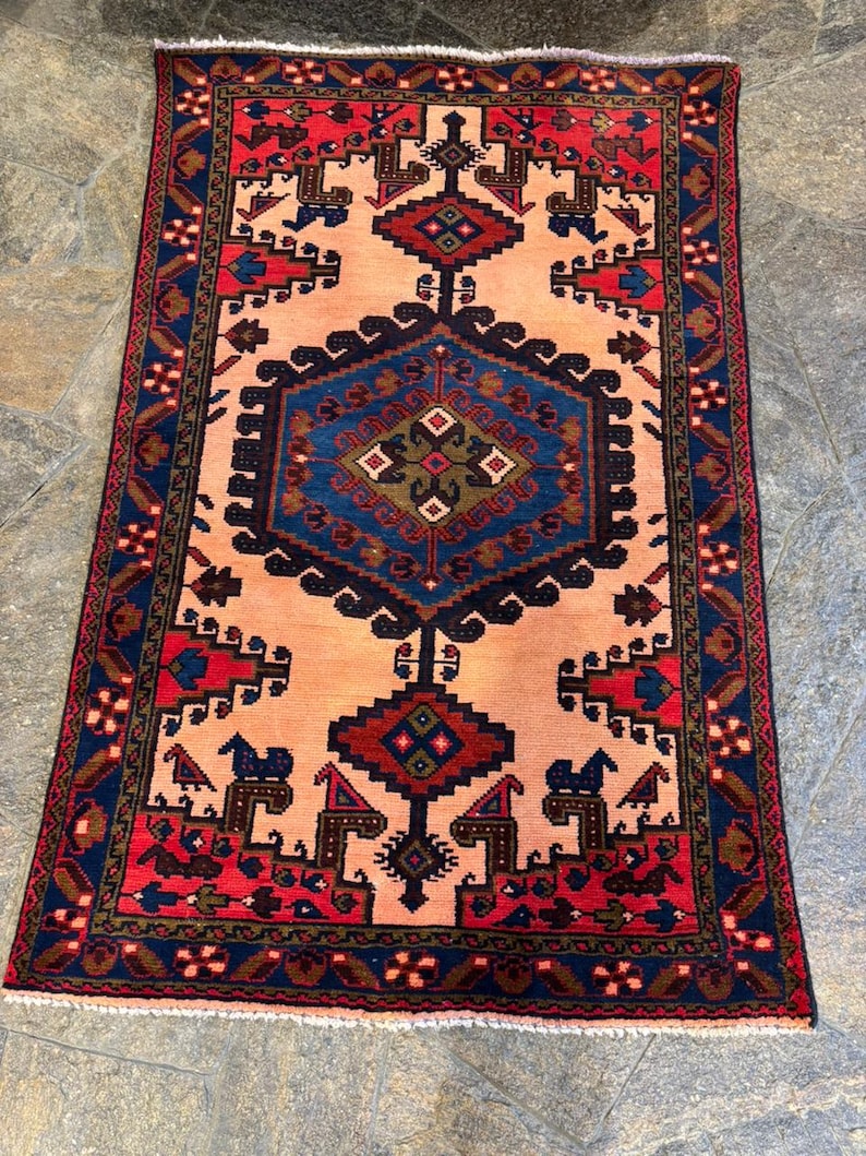May include: A rectangular rug with a central peach-colored field and intricate geometric patterns. The border is a deep navy blue with red and brown accents. The rug features repeating motifs of stylized shapes and figures, creating a traditional aesthetic.