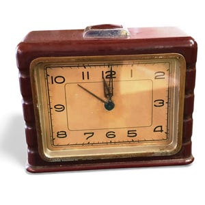 May include: A vintage maroon rectangular alarm clock with a gold-toned face and black numerals. The clock hands indicate approximately 11:50. The clock has a small handle on top and a gold-toned frame.