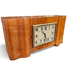 Op de afbeelding: Een vintage houten klok met een rechthoekige wijzerplaat en een goudkleurig frame. De wijzerplaat toont zwarte cijfers en wijzers tegen een crèmekleurige achtergrond. De houten behuizing heeft een gebogen ontwerp.