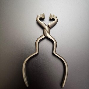 May include: Antique silver-toned sugar tongs with a twisted central design. The tongs feature decorative dragon-like heads at the gripping ends and curved handles. The object is set against a dark background, highlighting its intricate details and vintage aesthetic.