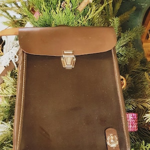 May include: A brown leather satchel with a flap closure and a metal clasp. The bag is rectangular with a rounded top and a strap closure on the bottom. The bag is set against a backdrop of evergreen branches.