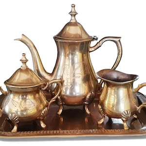 May include: A vintage gold-toned tea set, including a teapot, sugar bowl, and creamer, presented on a rectangular tray. The set features ornate detailing and is likely made of metal.
