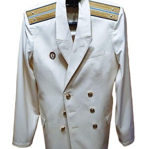 Puede incluir: Una blazer blanca de doble botonadura con botones dorados y charreteras. Las charreteras tienen ribetes dorados y rayas azul claro. Un pequeño pin está en la solapa izquierda. La chaqueta está colgada de una percha negra.