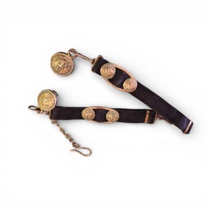 May include: Two antique black leather belts with gold-colored metal accents. Each belt features decorative round buttons and a clasp. One belt has a chain attached. The belts are likely vintage accessories.