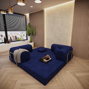 Könnte beinhalten: Ein modulares, marineblaues Sofa mit abgerundeten Arm- und Rückenlehnen. Das Sofa steht auf einem Holzboden, mit einem kleinen Tablett mit Blumen und einer Decke. Der Raum hat ein großes Fenster mit Stadtblick und eine dekorative Pflanze.