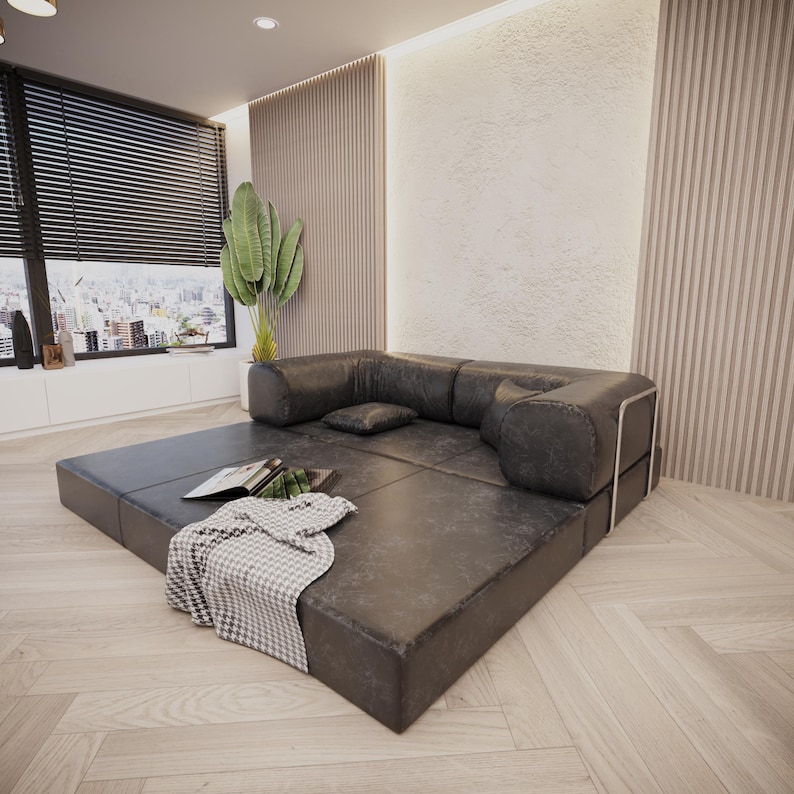 Puede incluir: Un sof&aacute; seccional moderno y de perfil bajo en cuero gris oscuro. El sof&aacute; tiene un dise&ntilde;o modular con una base cuadrada y reposabrazos redondeados. Una manta, almohadas y un libro est&aacute;n colocados en el sof&aacute;. La habitaci&oacute;n tiene grandes ventanas y una planta.