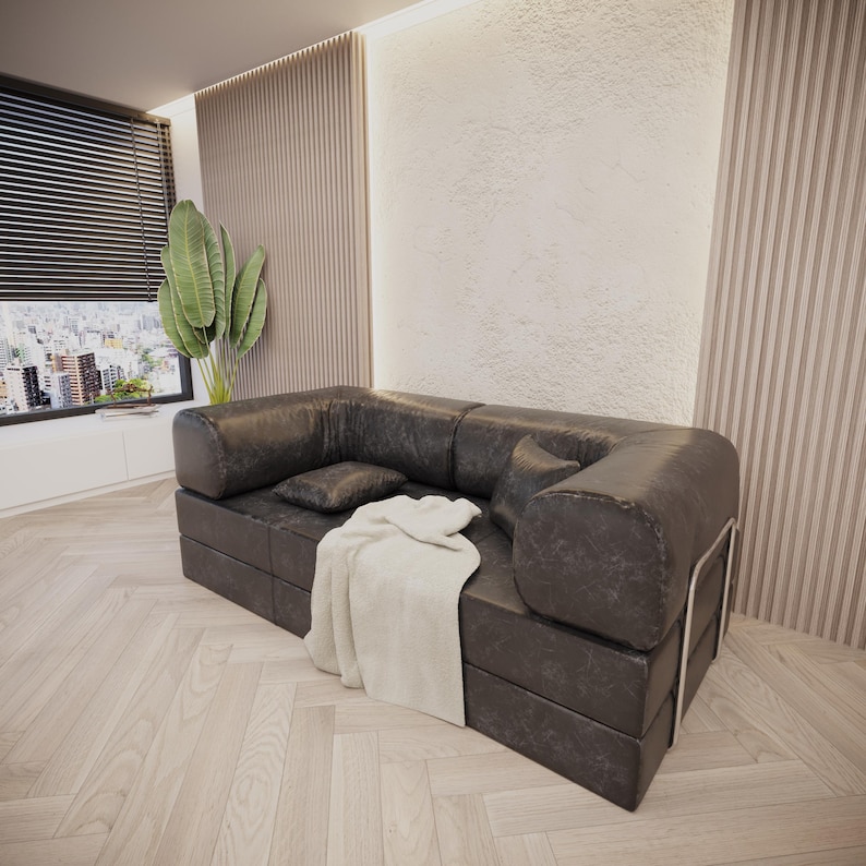 Puede incluir: Un sof&aacute; seccional de cuero gris oscuro con una manta blanca y dos cojines. El sof&aacute; est&aacute; en una sala de estar moderna con una ventana grande y pisos de madera. El sof&aacute; tiene un marco de metal.