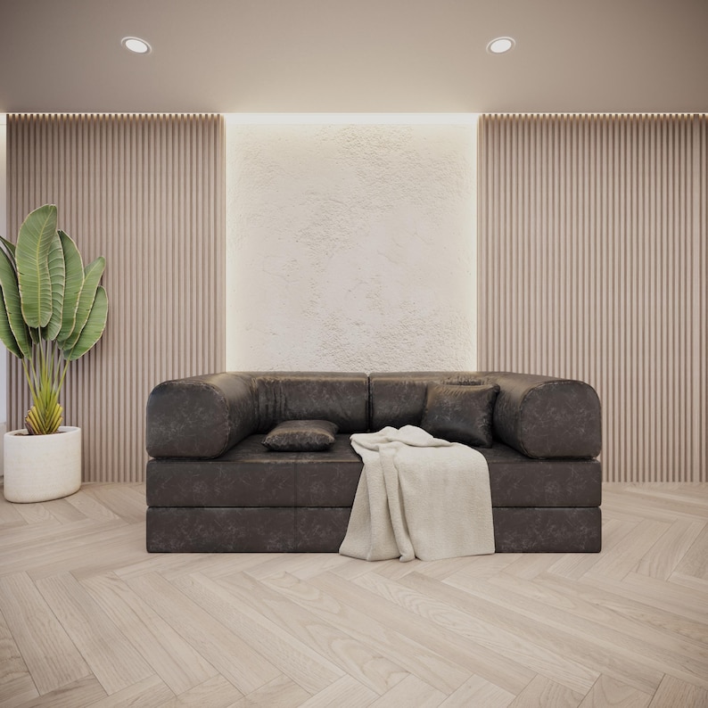 Puede incluir: Un sof&aacute; moderno de cuero marr&oacute;n oscuro con brazos y respaldo redondeados, con dos cojines cuadrados y un coj&iacute;n rectangular peque&ntilde;o. Una manta de color crema est&aacute; sobre el sof&aacute;. Una planta en maceta con hojas verdes grandes est&aacute; a la izquierda.