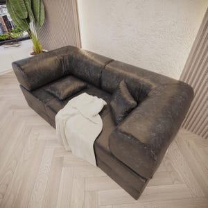 Puede incluir: Un sof&aacute; seccional de cuero gris oscuro con dos cojines a juego y una manta de color crema. El sof&aacute; tiene un aspecto texturizado y es de estilo moderno. La habitaci&oacute;n tiene piso de madera clara.