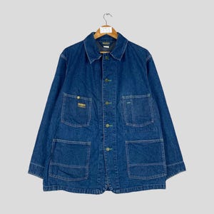 Puede incluir: Chaqueta vaquera azul vintage con m&uacute;ltiples bolsillos y cierres de botones. La chaqueta tiene cuello y est&aacute; colgada de una percha de madera. La chaqueta presenta un peque&ntilde;o parche con el logotipo en el bolsillo del pecho.