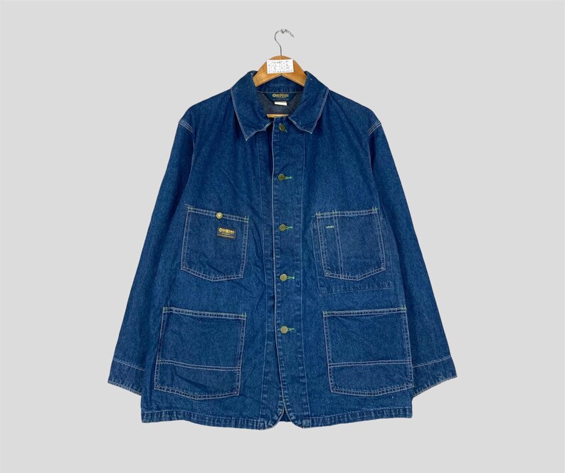 Puede incluir: Chaqueta vaquera azul vintage con m&uacute;ltiples bolsillos y cierres de botones. La chaqueta tiene cuello y est&aacute; colgada de una percha de madera. La chaqueta presenta un peque&ntilde;o parche con el logotipo en el bolsillo del pecho.