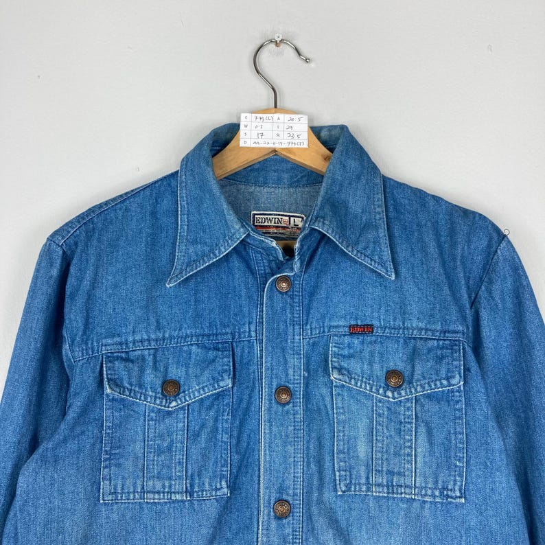 K&ouml;nnte beinhalten: Eine mittelblaue Jeansjacke mit Knopfleiste, zwei Brusttaschen und einem klassischen Kragen. Die Jacke tr&auml;gt das Markenetikett "EDWIN" und ein kleines rotes Etikett. Die Jacke h&auml;ngt an einem Holzkleiderb&uuml;gel.