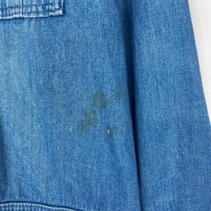 K&ouml;nnte beinhalten: Nahaufnahme eines hellblauen Jeanshemdes mit einer Taschendetail. Das Hemd hat mehrere dunkle Flecken auf der Vorderseite. Der Stoff scheint ein mittelschwerer Denim zu sein.