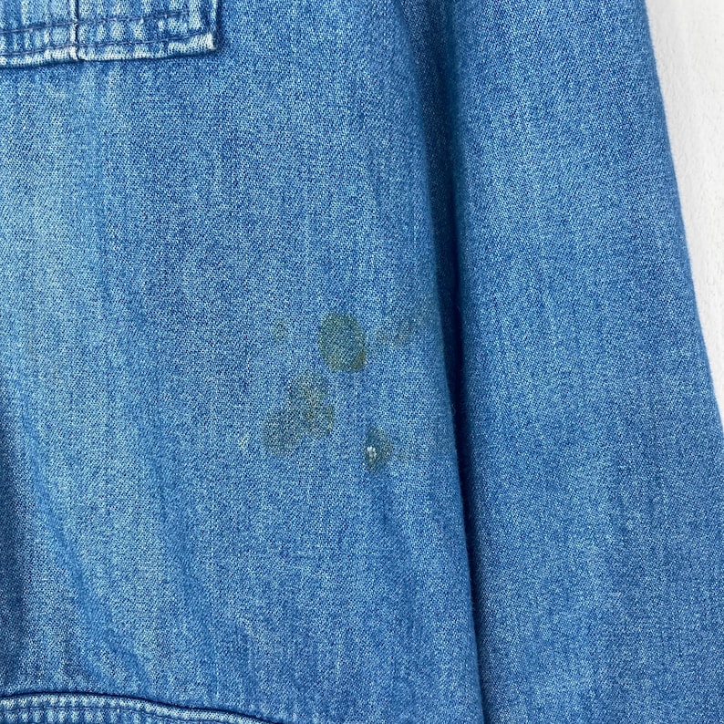 K&ouml;nnte beinhalten: Nahaufnahme eines hellblauen Jeanshemdes mit einer Taschendetail. Das Hemd hat mehrere dunkle Flecken auf der Vorderseite. Der Stoff scheint ein mittelschwerer Denim zu sein.