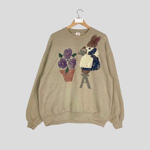 May include: Tan sweatshirt with a floral and character design. The design includes purple flowers in a pot, a character watering the flowers, and the letter "A". The sweatshirt is hanging on a wooden hanger.