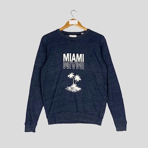 Puede incluir: Una sudadera azul marino con la palabra "MIAMI" en letras blancas, con un efecto espejo debajo. Debajo del texto hay un gráfico blanco de dos palmeras en una pequeña isla.