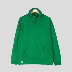 Puede incluir: Una sudadera verde brillante con cuello chal y bolsillo delantero. La sudadera tiene cierre de tres botones y un pequeño parche con el logo cerca del dobladillo. Cuelga de una percha de madera.