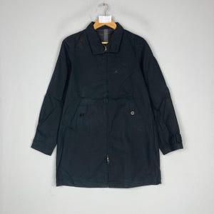 Puede incluir: Un abrigo largo negro con cuello, cremallera y cierres de botón. El abrigo tiene mangas largas y un dobladillo recto. El abrigo está colgado de una percha sobre un fondo gris claro.